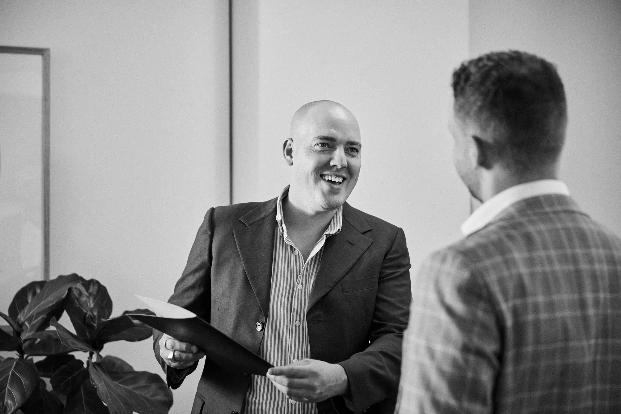 A smiling man in a suit talking to another man whose back it to us.